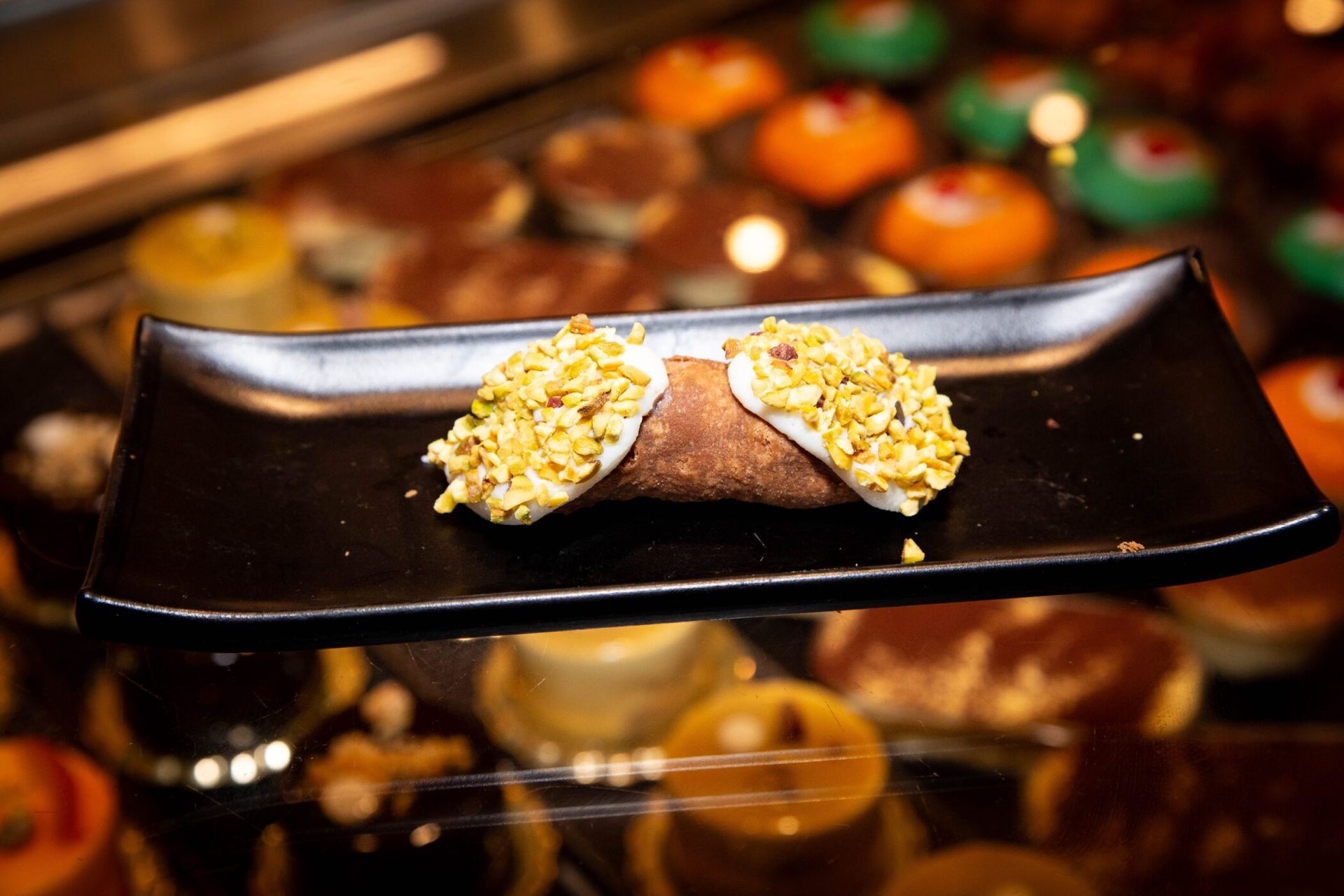 Cannoli on a black plate, with pistachio garnish, in a bakery display case.