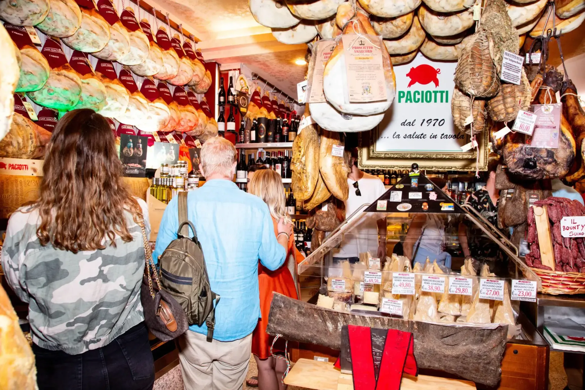 People browsing cured meats at a deli counter, surrounded by hanging products.