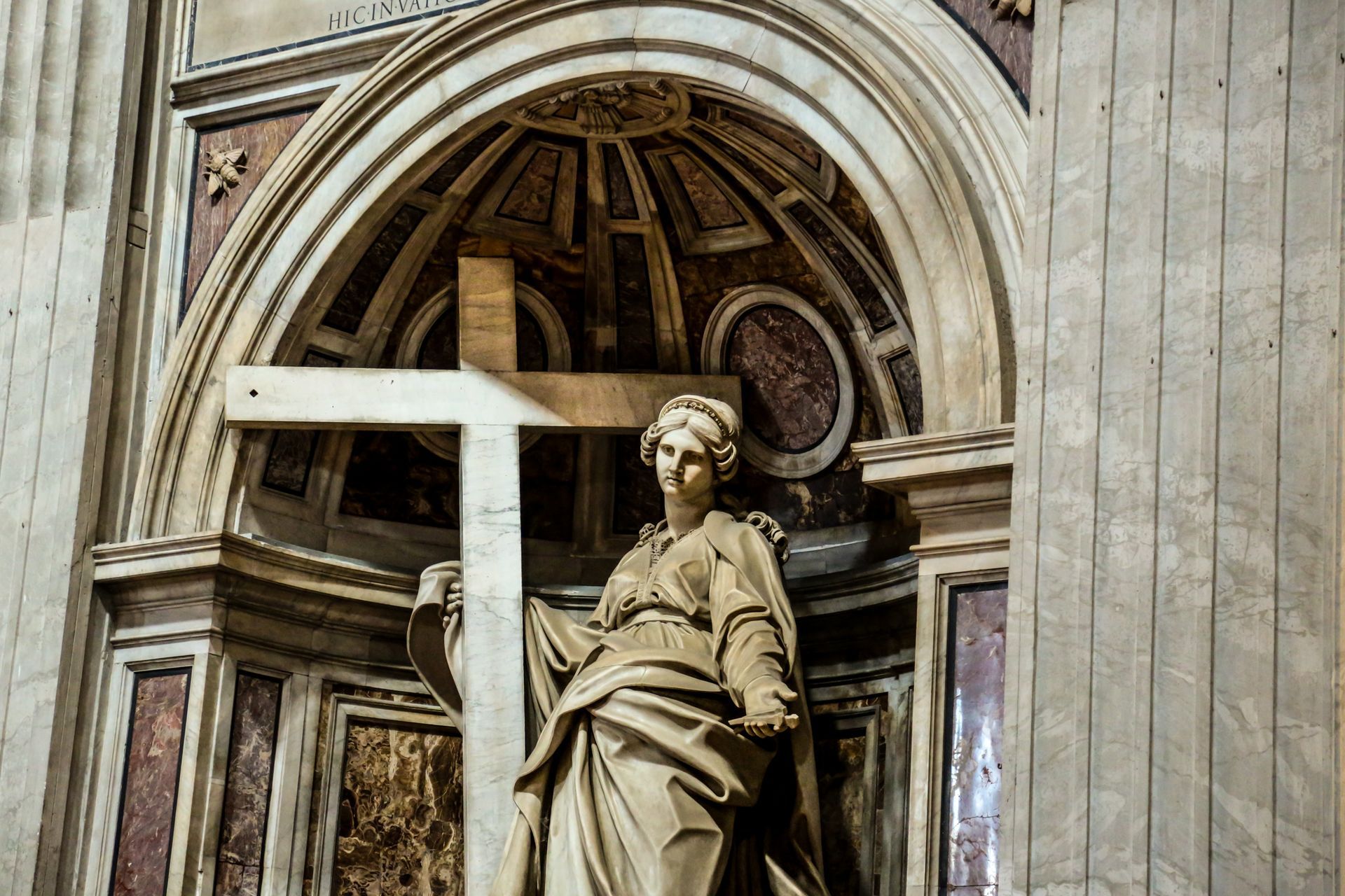 Statue of a woman holding a cross inside a church, marble, beneath an arched alcove with ornate details.