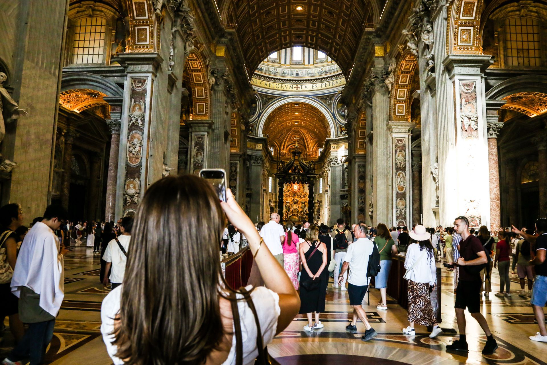 Woman takes a photo inside St. Peter's Basilica, Rome, with other tourists. Ornate gold and marble interior.