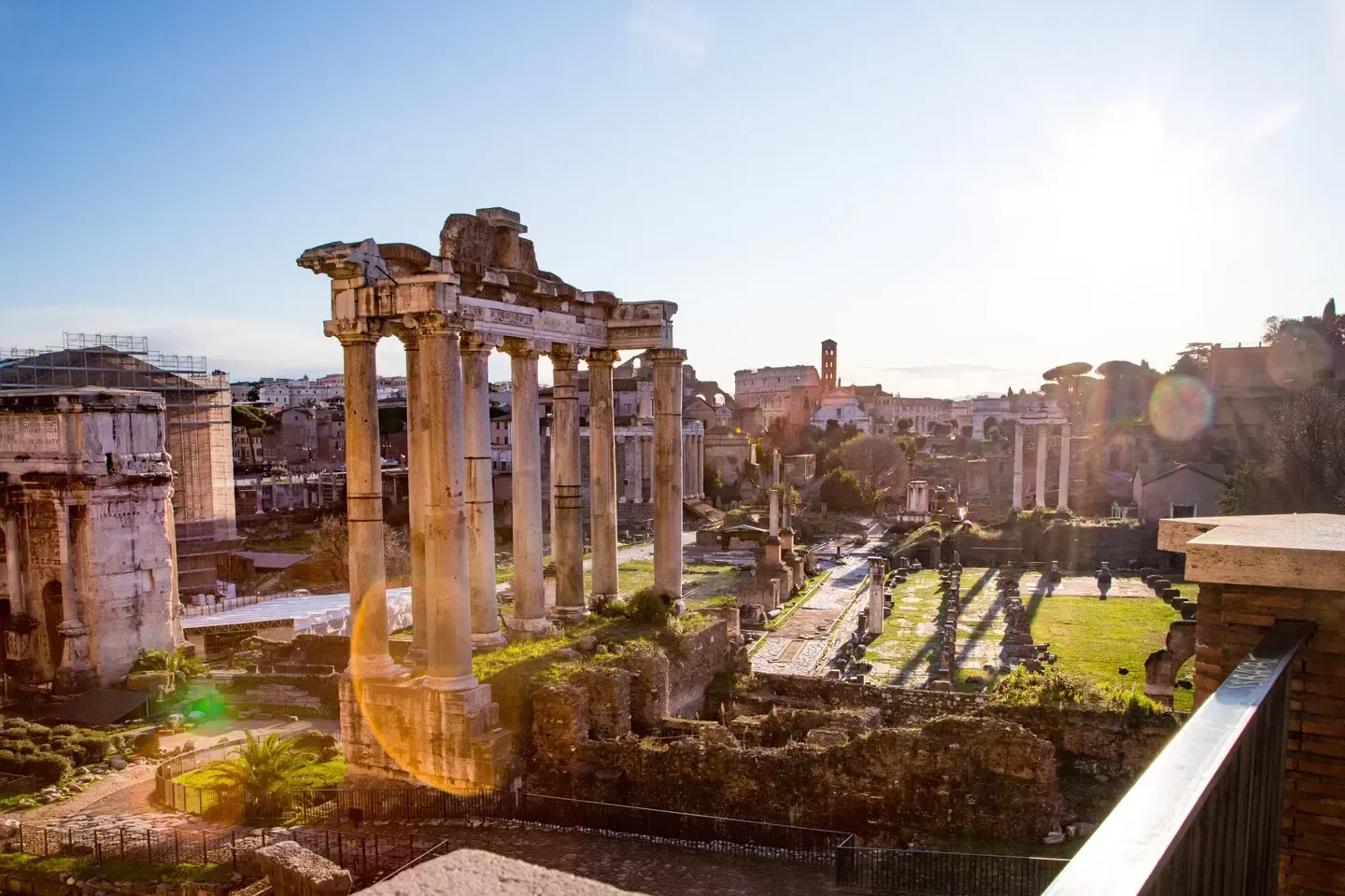 Ancient Roman ruins bathed in sunlight. Columns and arches frame a grassy area.