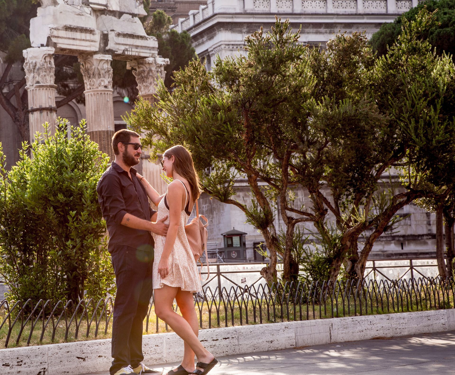 Romantic couple with ancient Roman ruins with tall columns, backlit by the sun, set behind a metal fence and red flowers.
