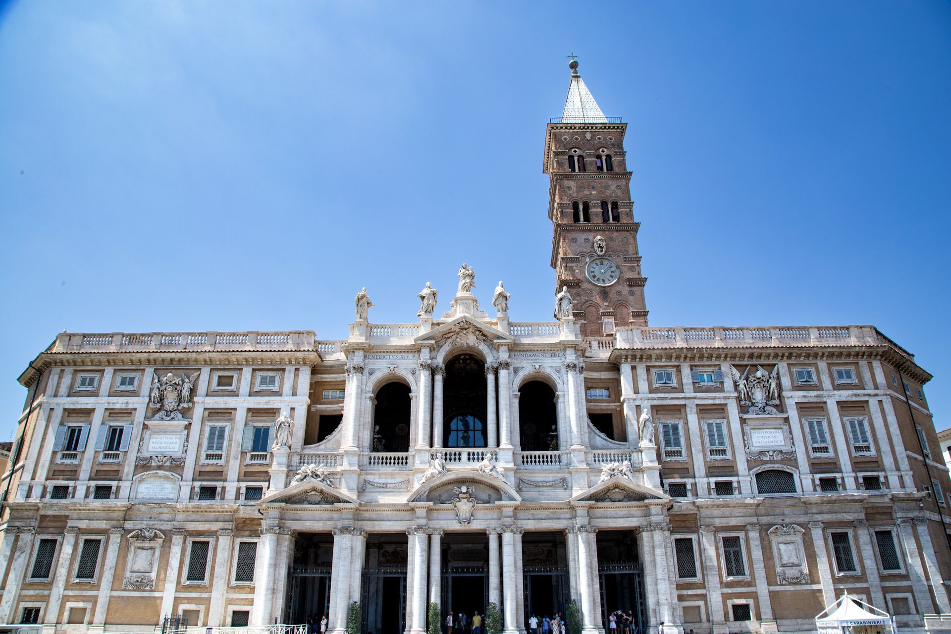 Stone bridge with statues over a river in Rome, Italy. Buildings and trees line the banks under a cloudy sky.