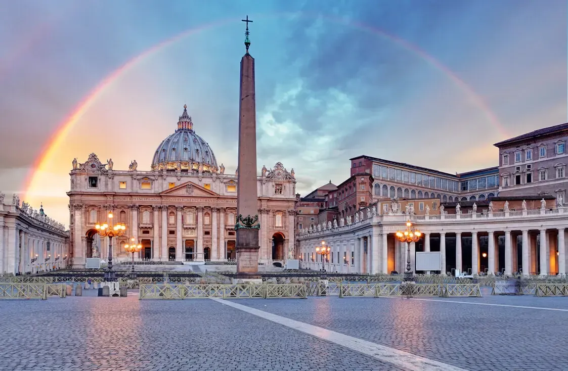 St. Peter's Basilica with rainbow in the sky. Cobblestone square, tall obelisk, buildings with lights.