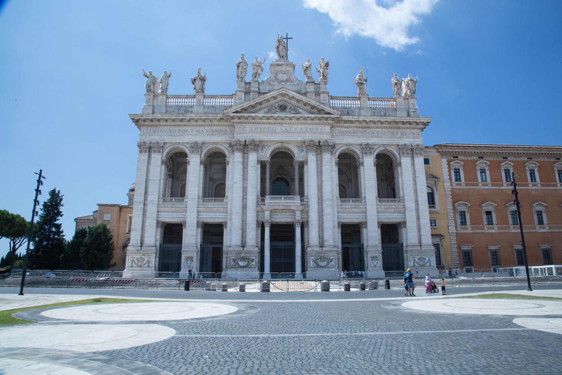Stone bridge with statues over a river in Rome, Italy. Buildings and trees line the banks under a cloudy sky.