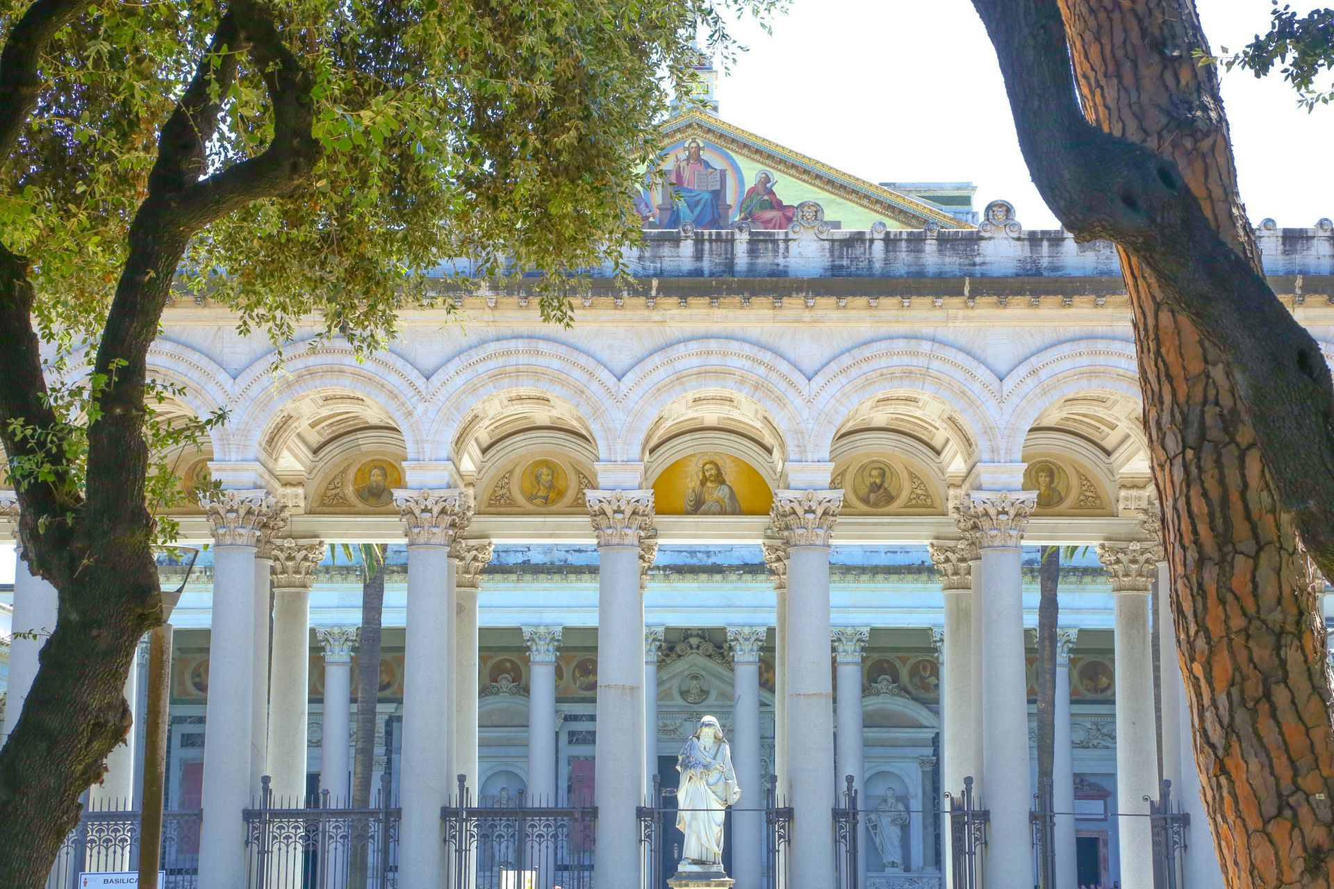 Stone bridge with statues over a river in Rome, Italy. Buildings and trees line the banks under a cloudy sky.