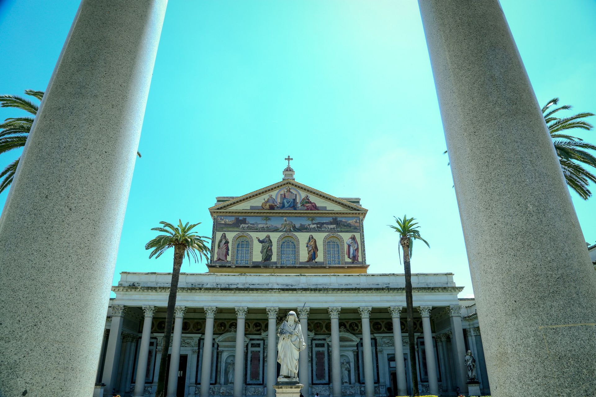 Stone bridge with statues over a river in Rome, Italy. Buildings and trees line the banks under a cloudy sky.