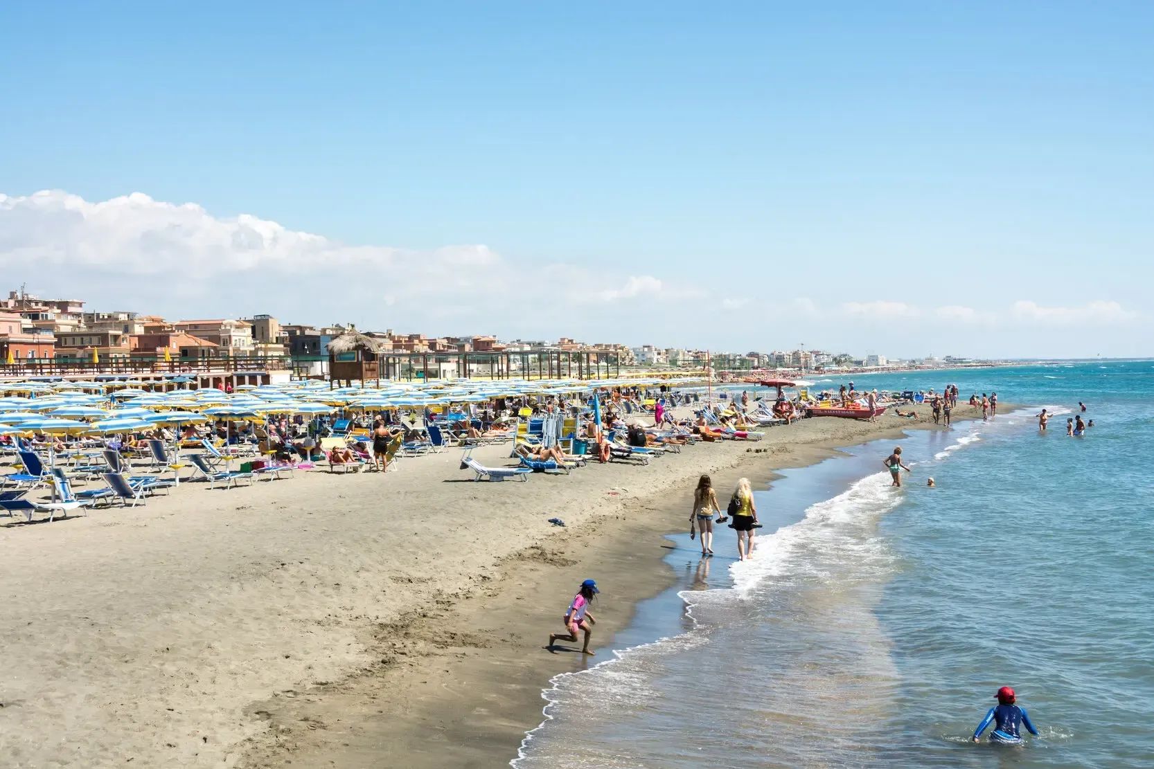 Beach scene: People relax and play along the shore, lined with umbrellas and buildings under a bright blue sky.