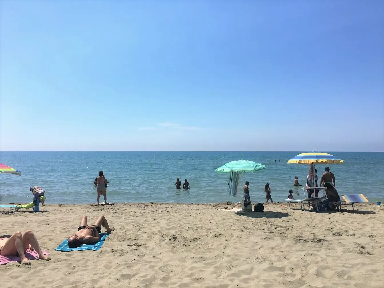 Beach scene with people swimming, sunbathing, and relaxing under umbrellas on a sunny day.