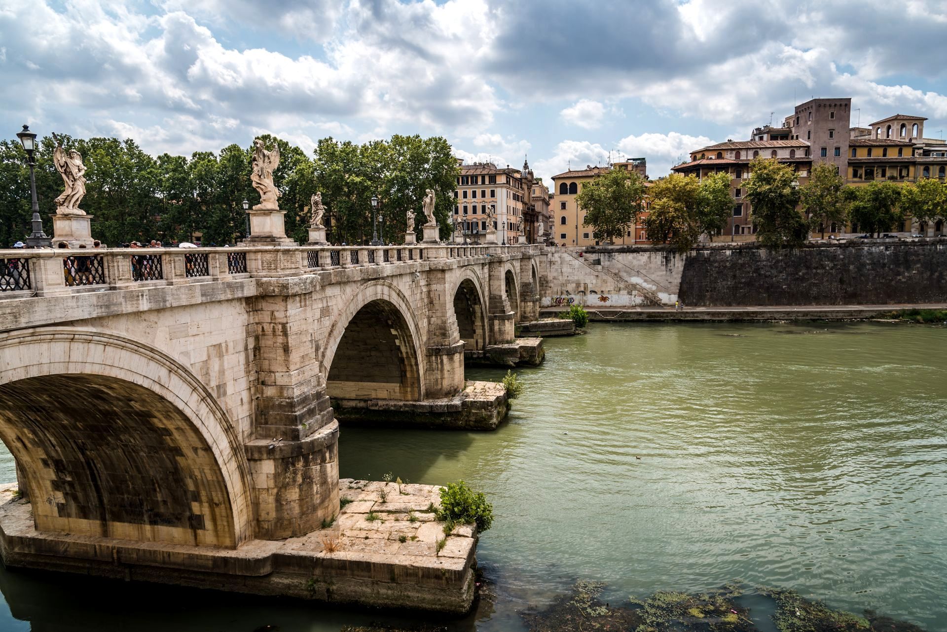 Stone bridge with arched spans over a river in Rome. Buildings and statues are visible.