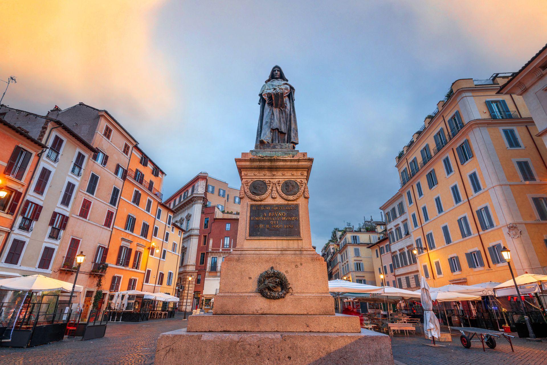 Statue in a plaza with colorful buildings and outdoor seating at dusk.