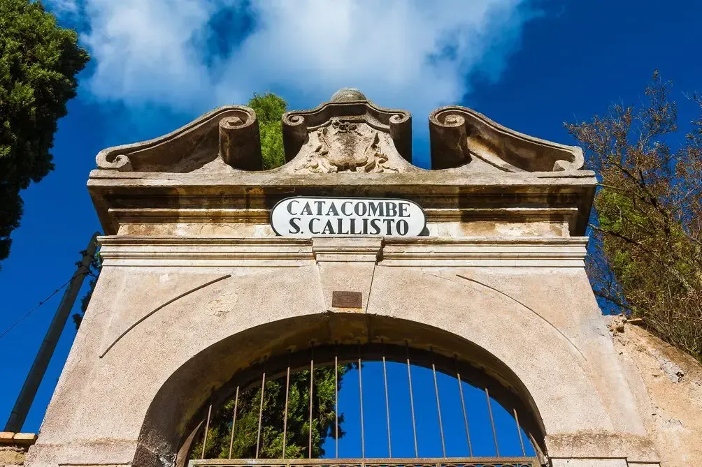 Stone archway entrance to the Catacombs of St. Callixtus with sign, under a blue sky.