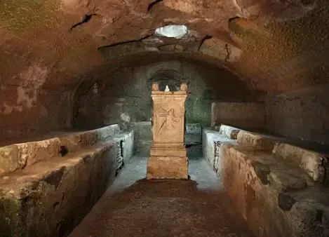 Stone-lined Roman tomb with alcoves and a central pillar. Dim lighting in an underground setting.