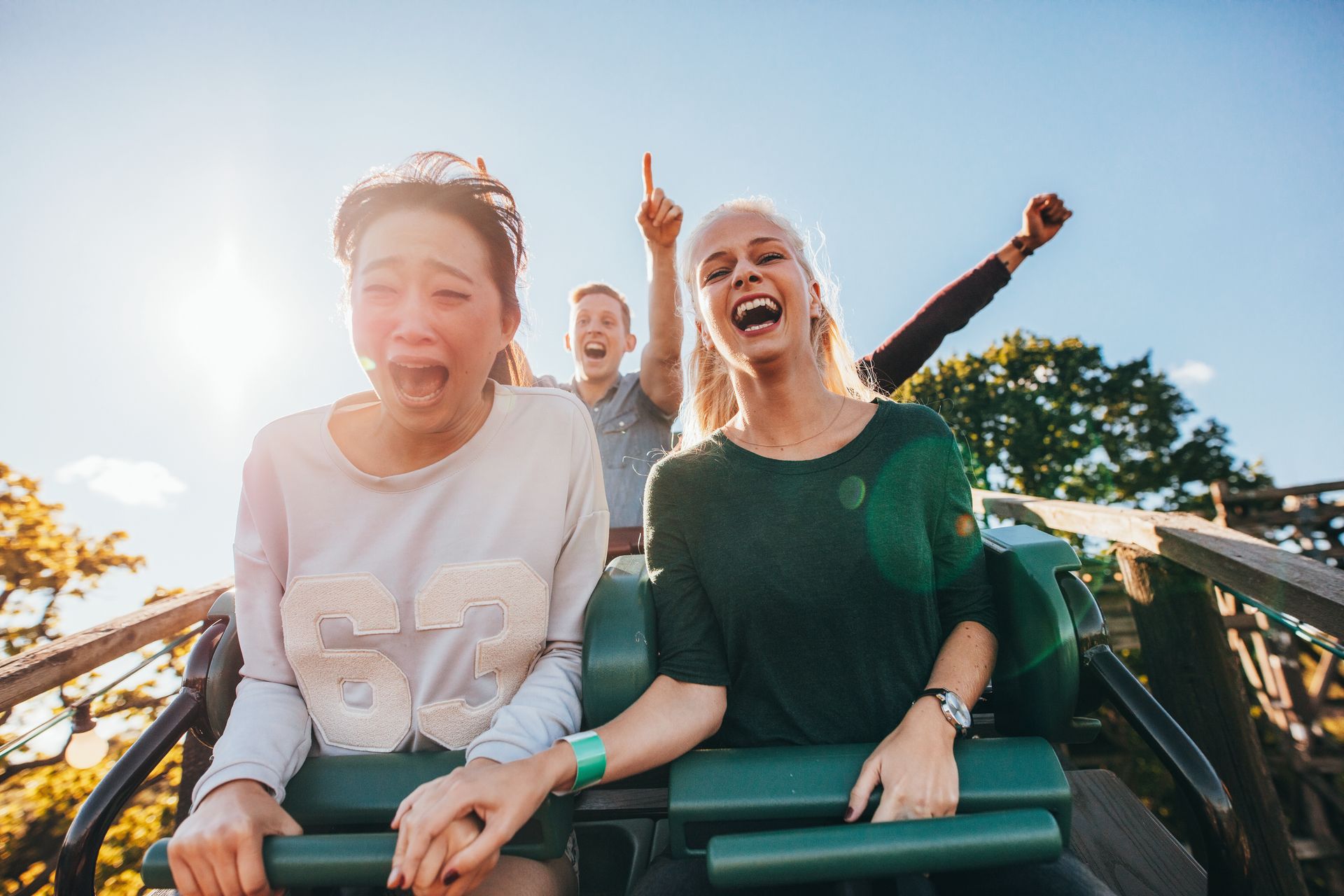 People on a roller coaster; one person screams, another laughs, a third raises a hand. Sunny, outdoor setting.