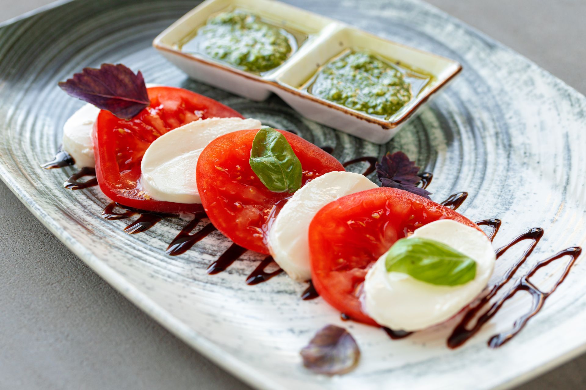 Caprese salad: tomato and mozzarella slices with pesto, balsamic glaze, and basil on a decorative plate.