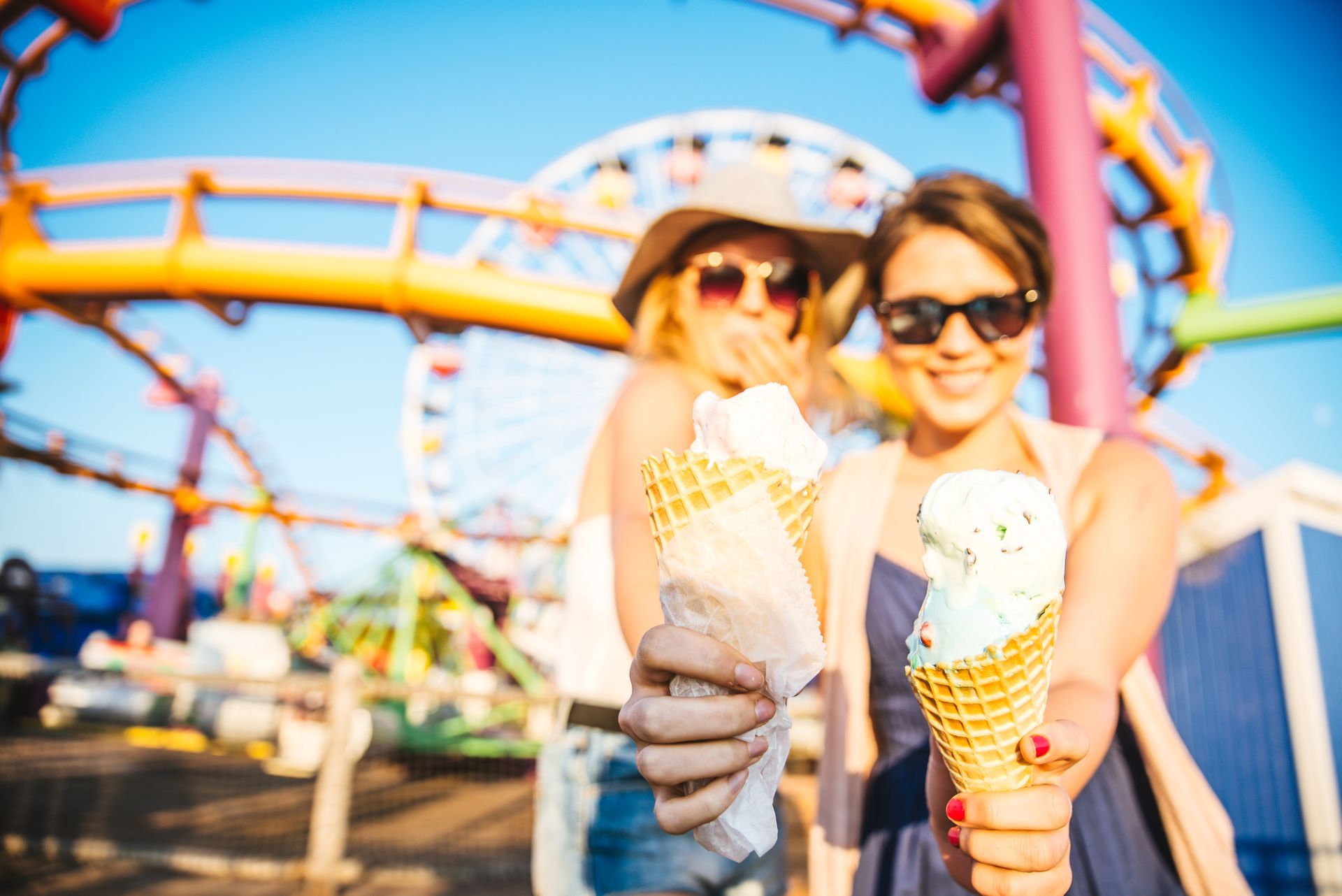 Two women holding ice cream cones at an amusement park; roller coaster and Ferris wheel in background.