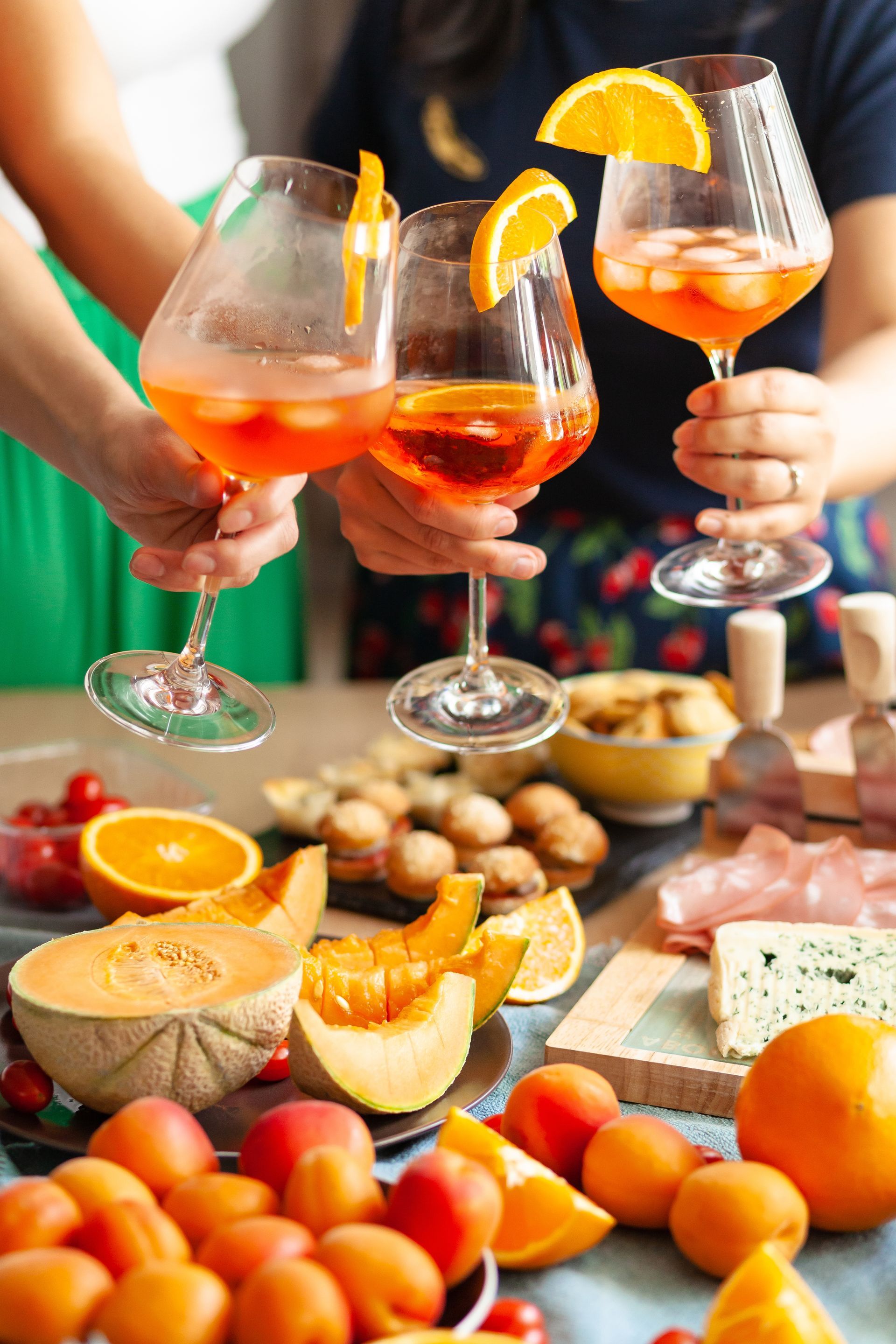 People toasting with Aperol spritzes, surrounded by fruit and snacks.