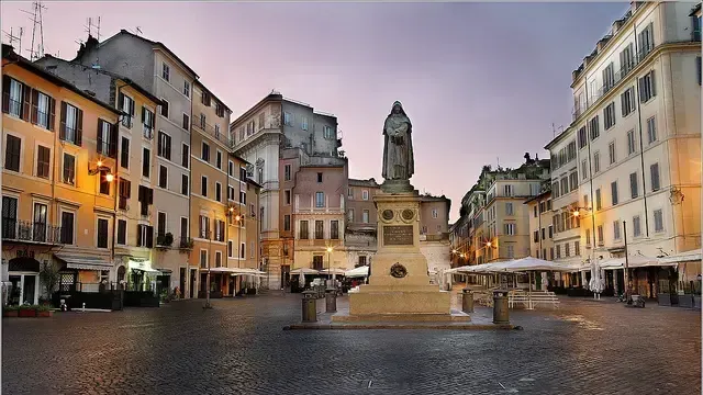 A square in Rome, Italy, with historic buildings, cobblestones, a monument, and outdoor dining at dusk.