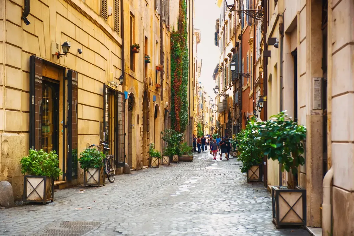 Narrow cobblestone street in Rome lined with buildings and potted plants.