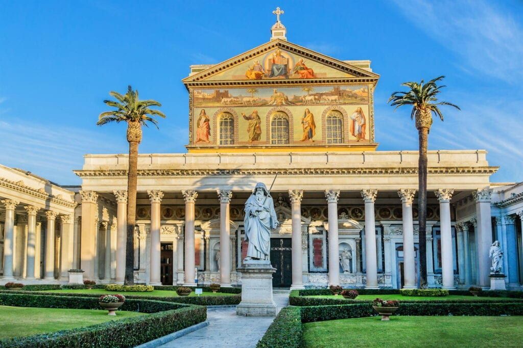 Basilica di San Paolo fuori le Mura, Rome, Italy, with columns, mosaic facade, and statue in a garden, under a blue sky.