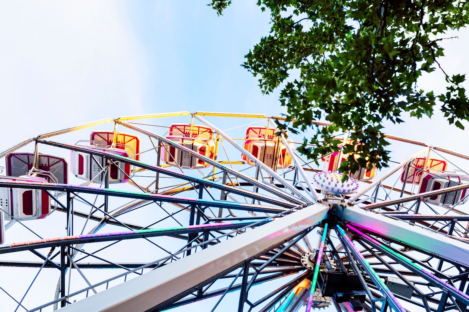 Ferris wheel against a cloudy blue sky, with illuminated colorful lights and green tree leaves.