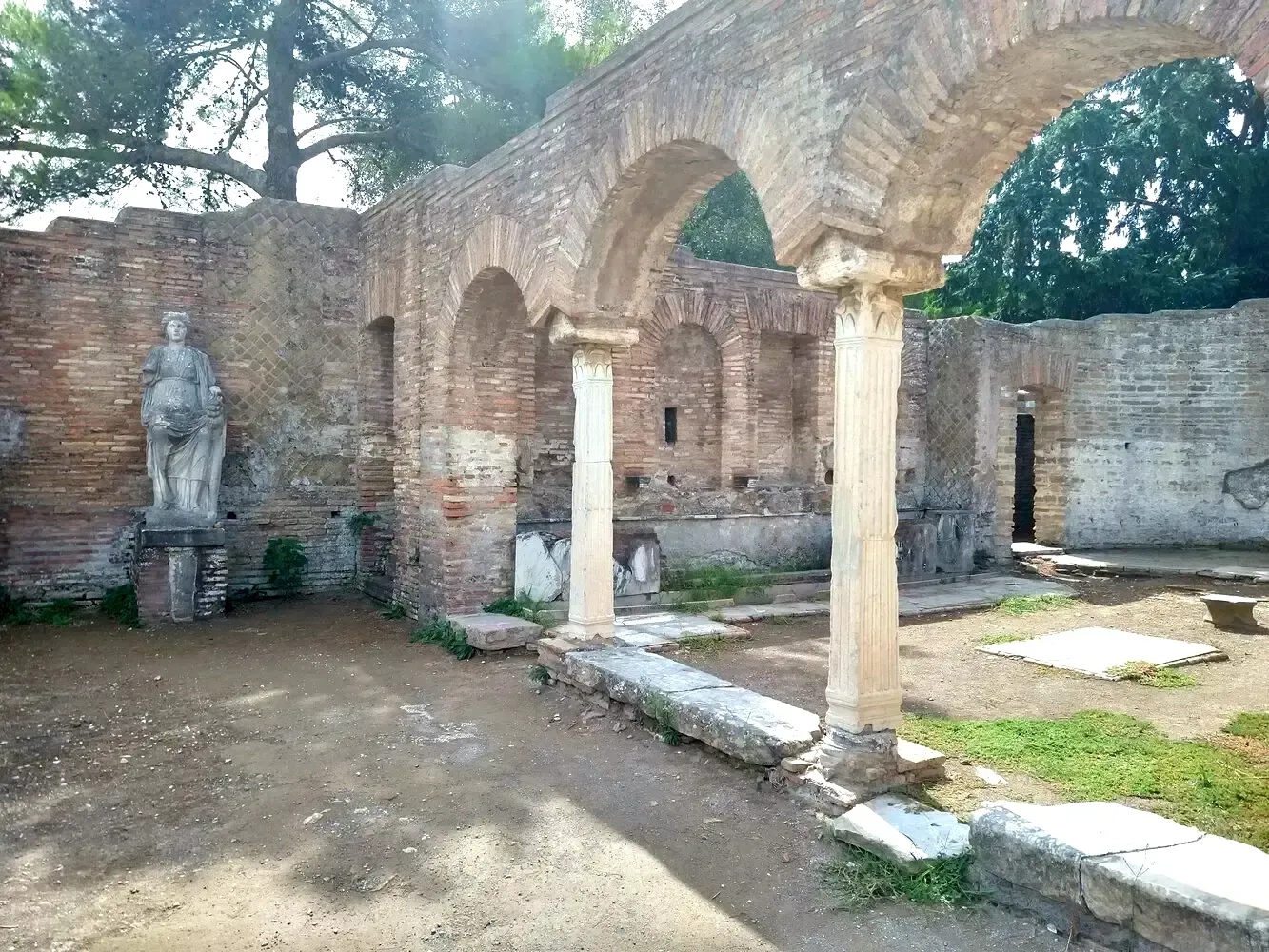 Ruins of an ancient structure with brick arches, columns, and a statue, outdoors.