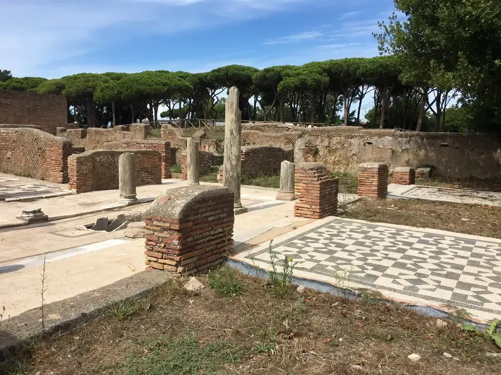 Ancient Roman ruins with brick walls, columns, mosaic floors, and trees in the background under a blue sky.