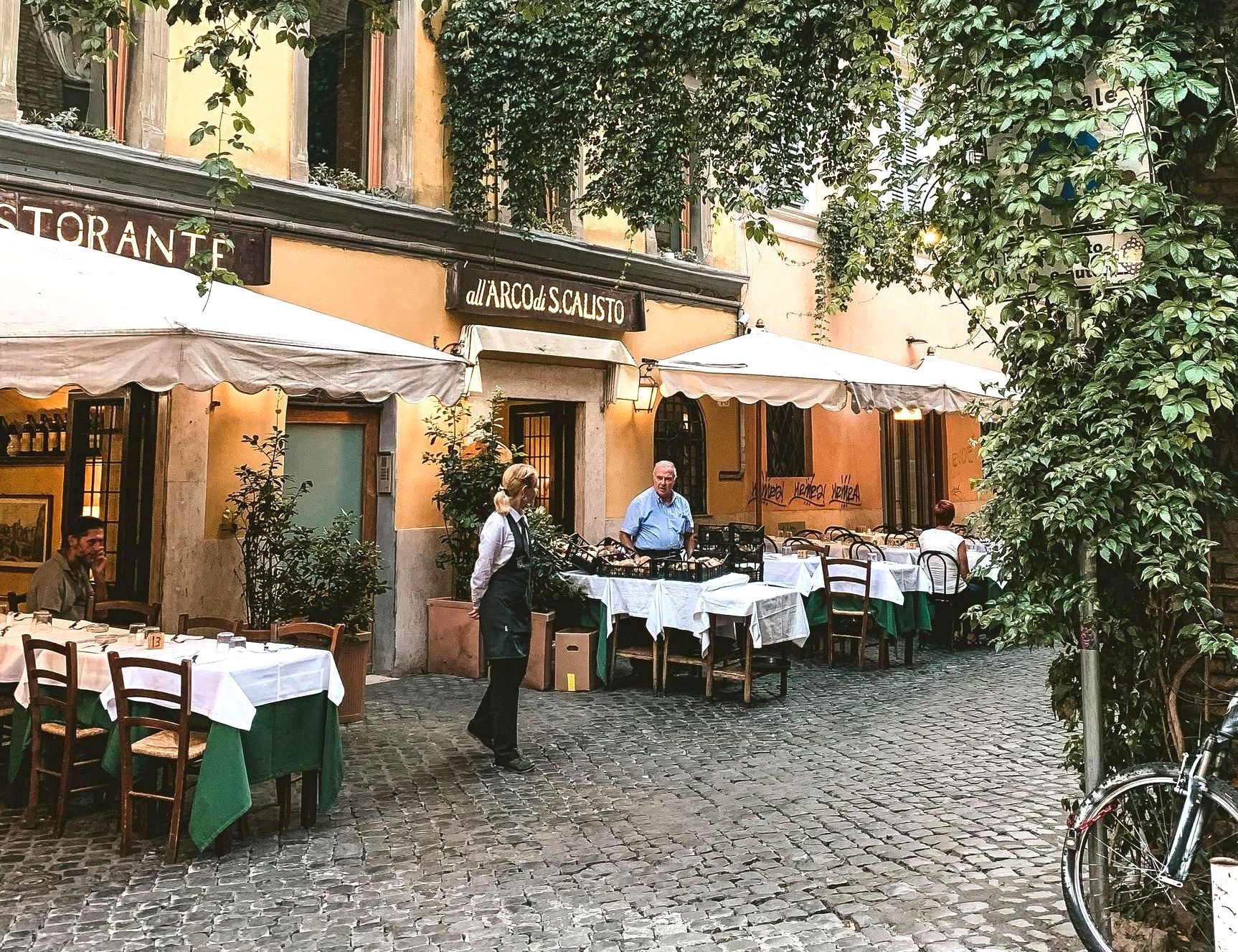 Restaurant with outdoor seating under awnings. Person stands near tables set with white tablecloths and green accents.