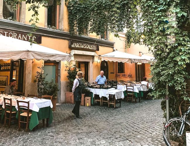 Restaurant with outdoor seating under awnings. Person stands near tables set with white tablecloths and green accents.