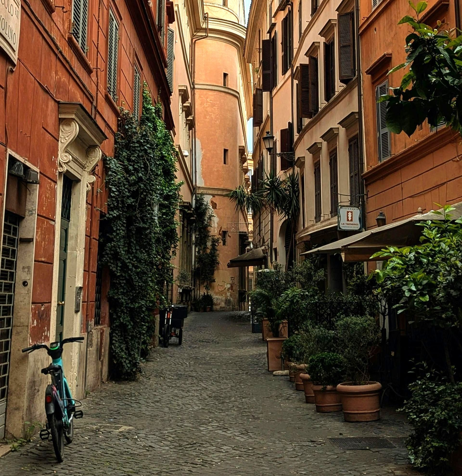 Cobblestone alley in Rome, Italy, lined with buildings in orange and brown tones; potted plants and a bicycle.