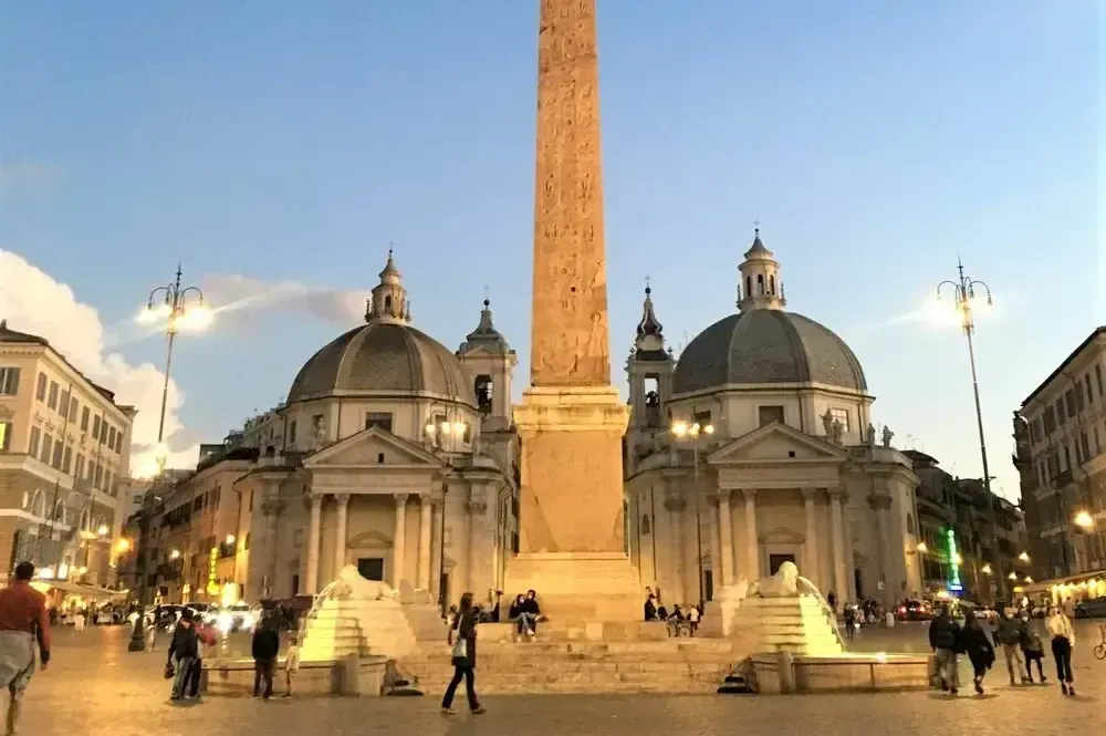 Obelisk and twin churches in Piazza del Popolo, Rome, at dusk. Buildings are lit, people are present.