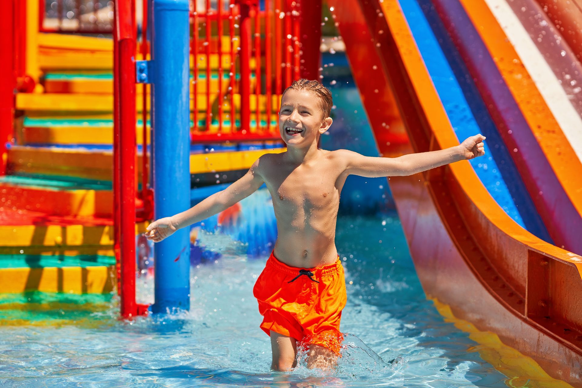Boy in orange swim trunks, smiling with arms outstretched in a water park, near a colorful waterslide.