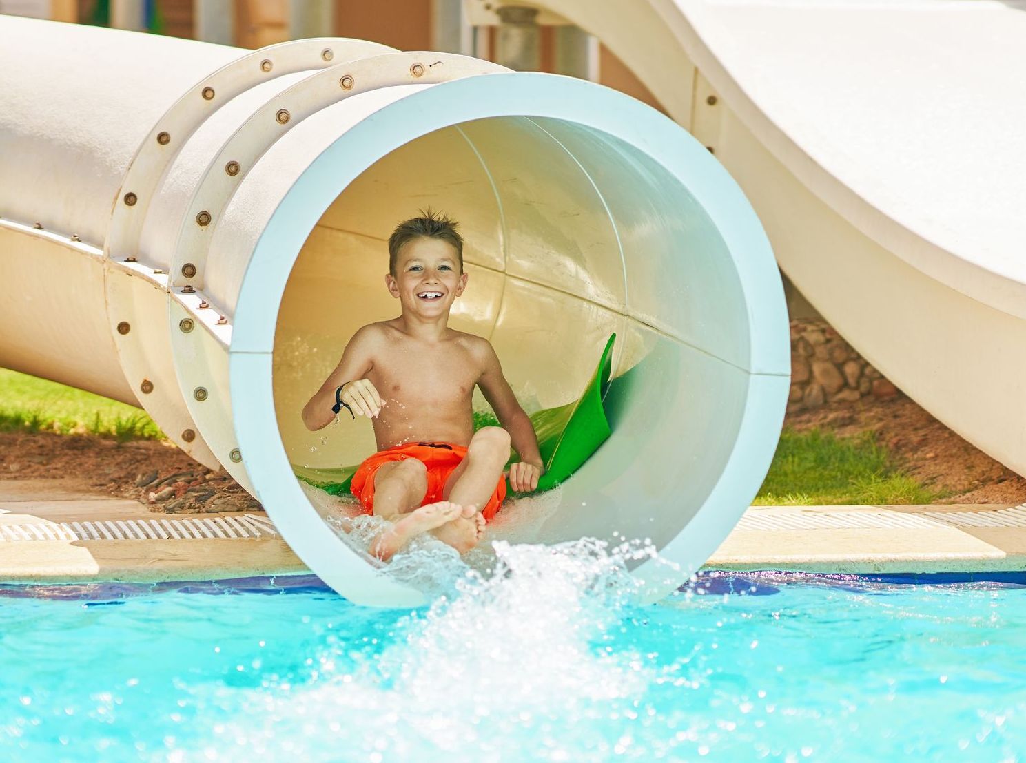 Boy exiting a water slide into a pool, smiling, with water splashing.