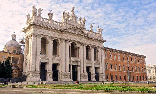 Exterior view of the Papal Archbasilica of St. John Lateran, Rome, with white facade, statues, and red building on the side.