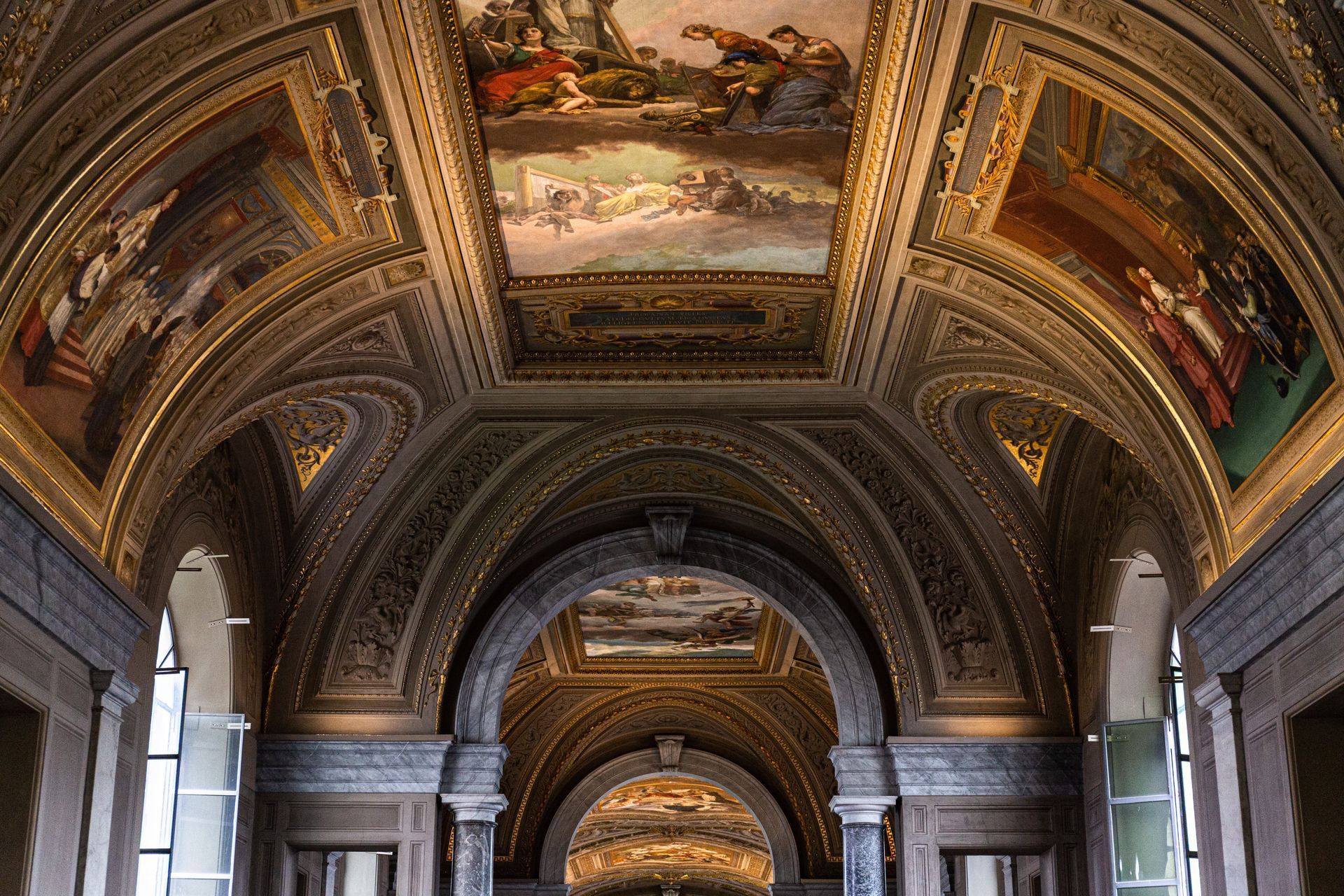 Ornate vaulted ceiling with painted panels, archways, and detailed gold trim in a building interior.