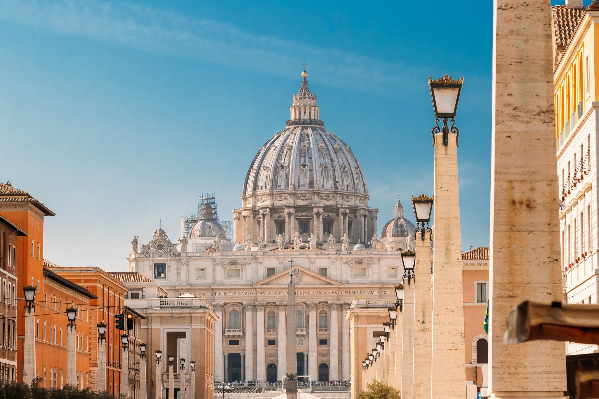 St. Peter's Basilica, Rome, seen from a street lined with columns and streetlights; blue sky above.
