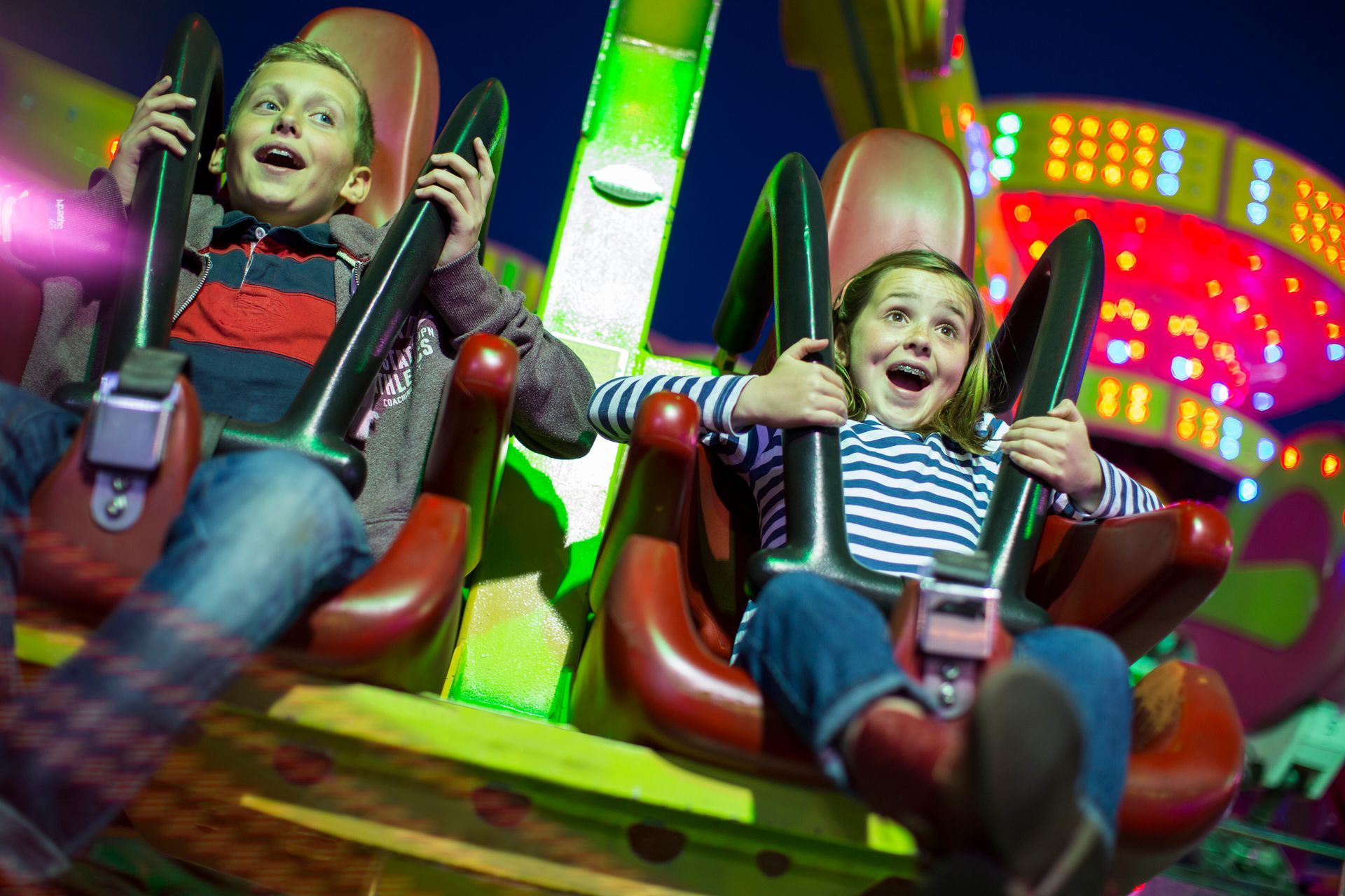 Two children on amusement park ride, smiling with excitement, arms up. Green and pink lights in background.