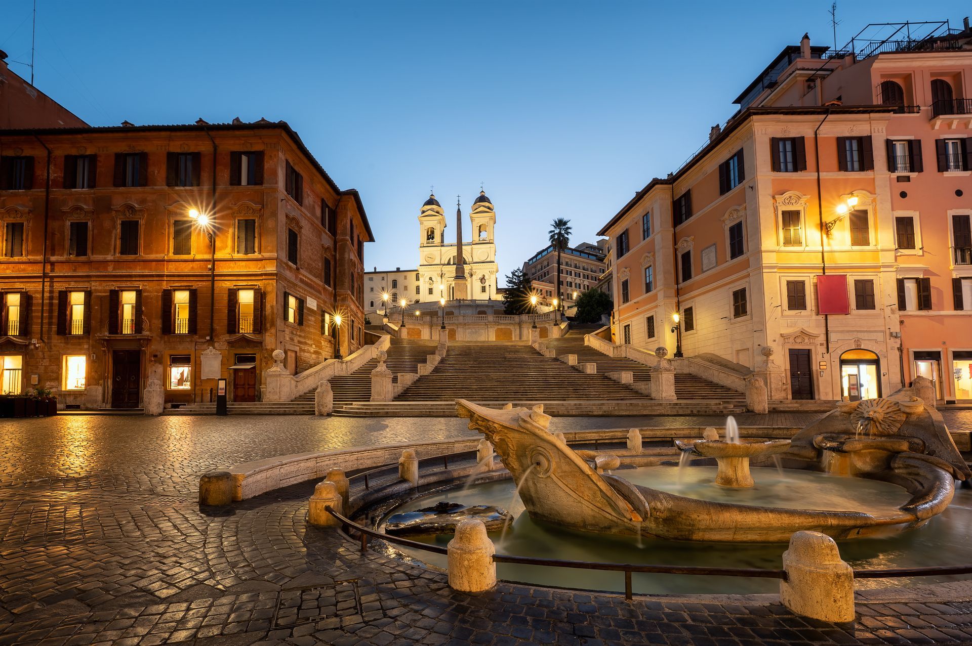 Spanish Steps and fountain in Rome at dusk. Buildings frame the stairs leading to a church.