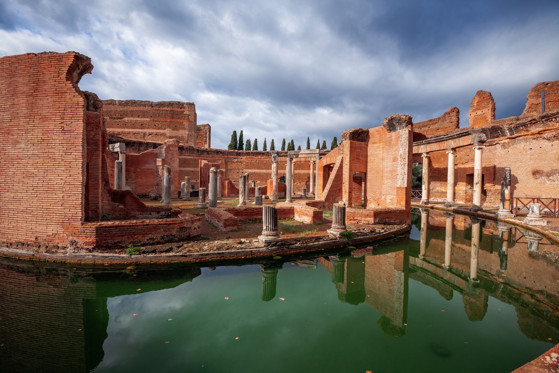 Ancient brick ruins with white columns reflected in still green water under a cloudy sky.