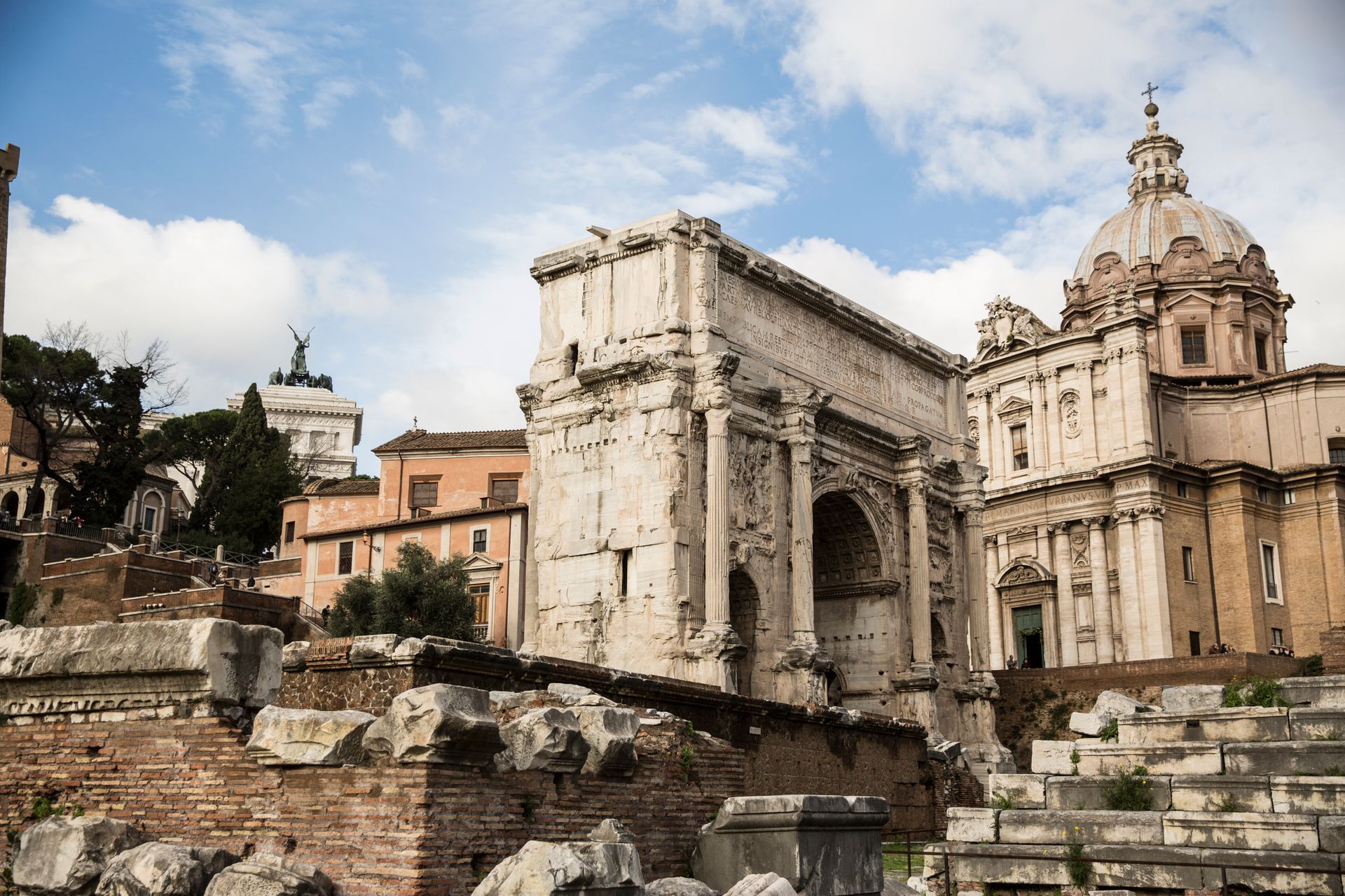 Ruins of ancient Roman buildings with a triumphal arch, under a cloudy sky.
