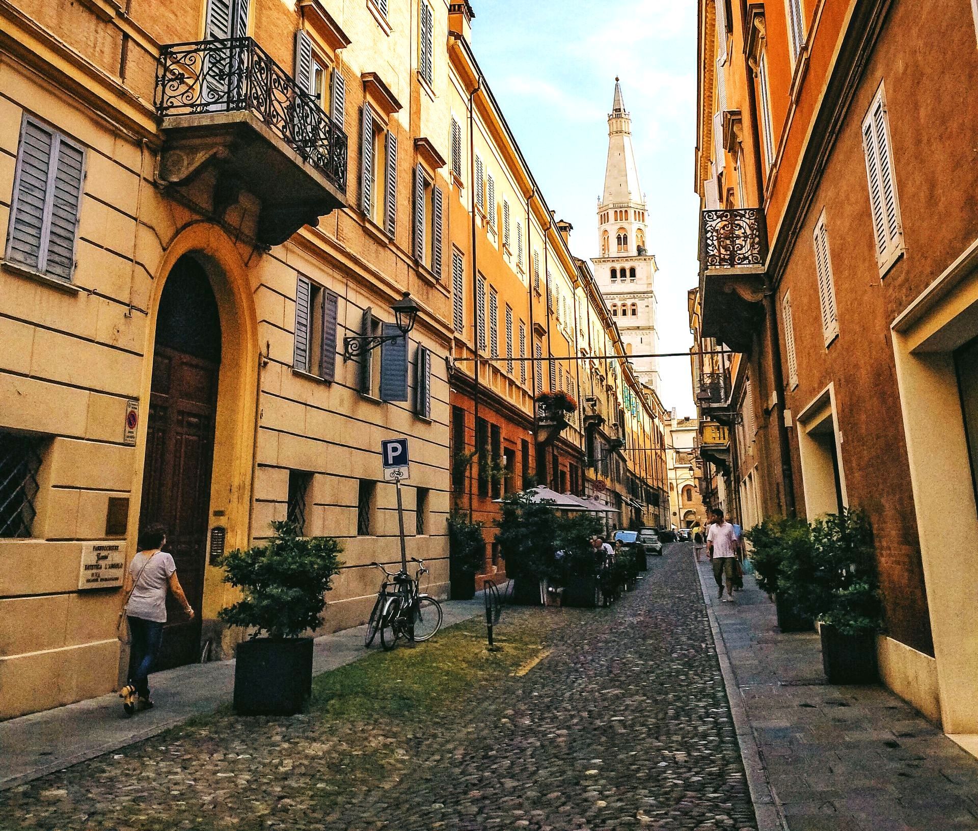 Cobblestone street in Modena, Italy, lined with buildings; the Duomo bell tower is visible at the end of the street.