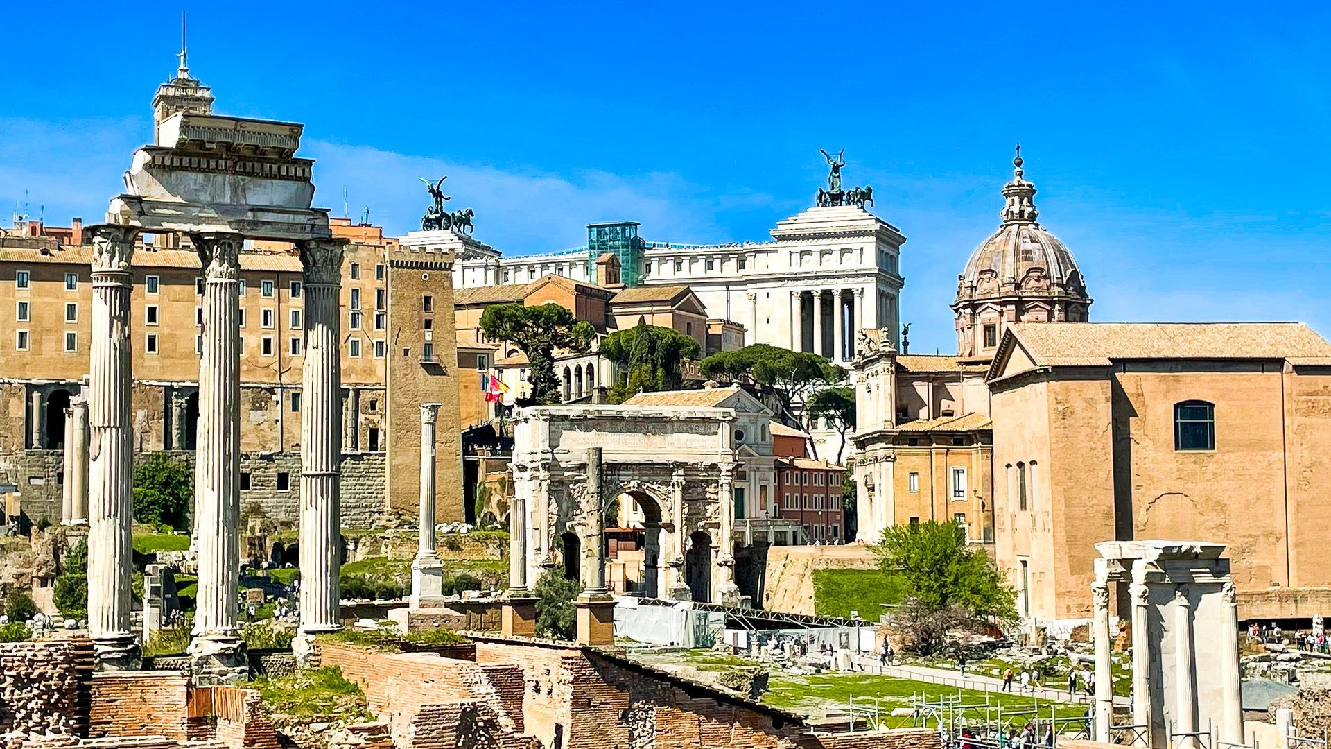 Ruins and buildings of the Roman Forum, with columns, archways, and a dome under a clear blue sky.