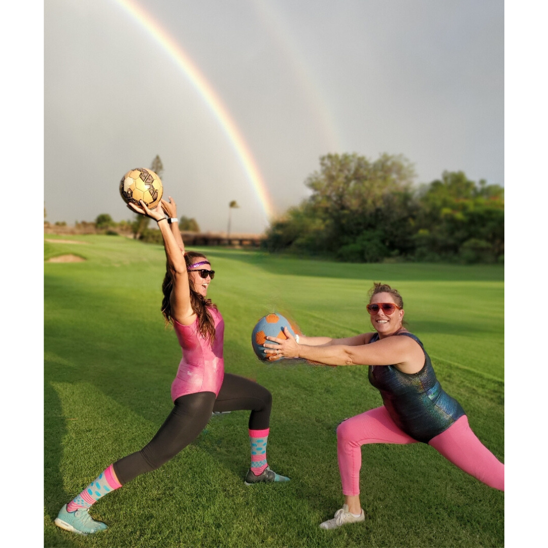 Two women are playing with a ball in a field with a rainbow in the background