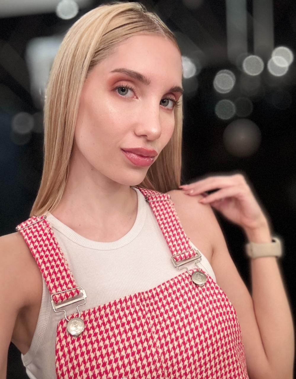 Mujer rubia con mono rojo y blanco, camisa blanca, mano apoyada en el hombro, mirando a la cámara.