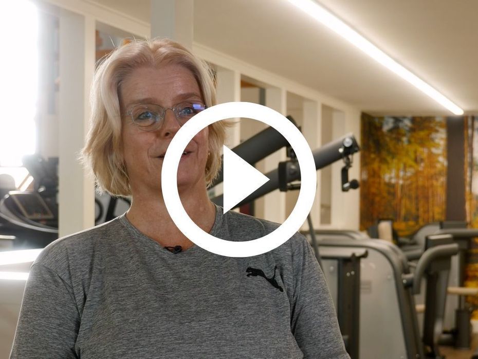 Een persoon in een grijs shirt met lange mouwen en een speelknop erop, staat in een sportschool met fitnessapparatuur.