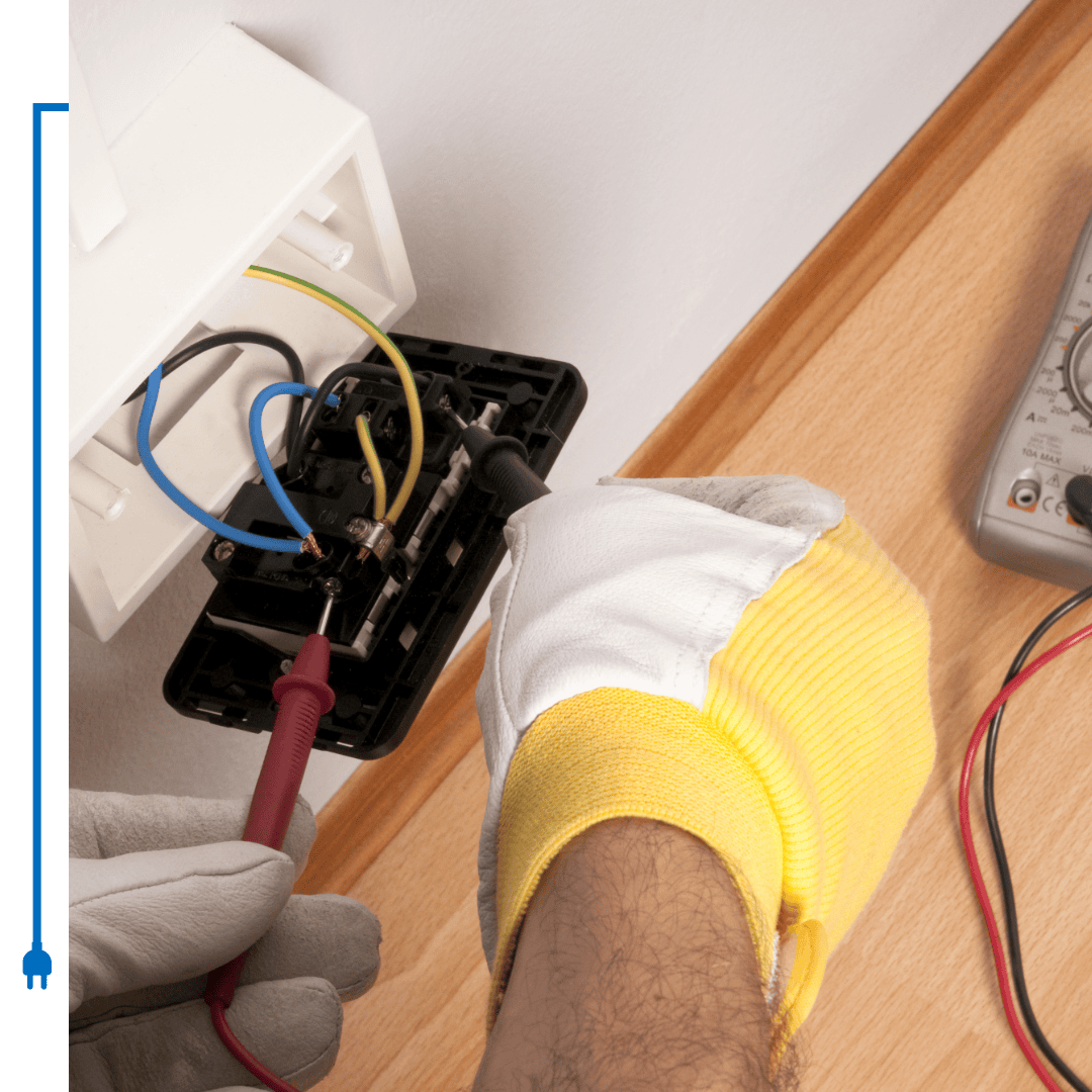 Electrician using a multimeter to test a wall outlet, wearing gloves, indoors.