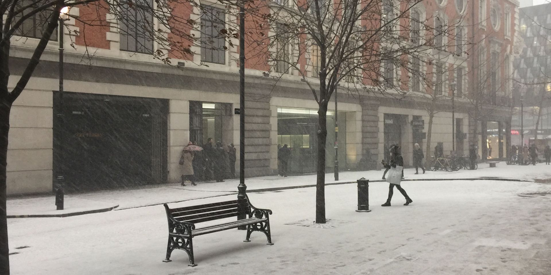 Snowy street scene with people walking past buildings. A bench sits in the foreground.