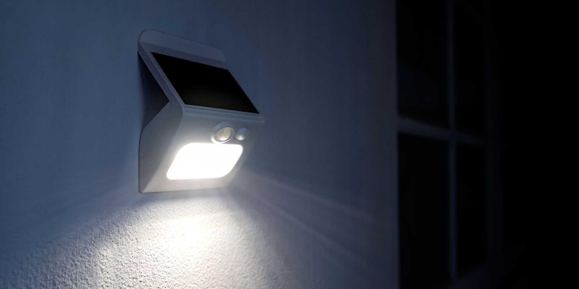 A white solar-powered light fixture with a bright light illuminating a wall in a dark setting.
