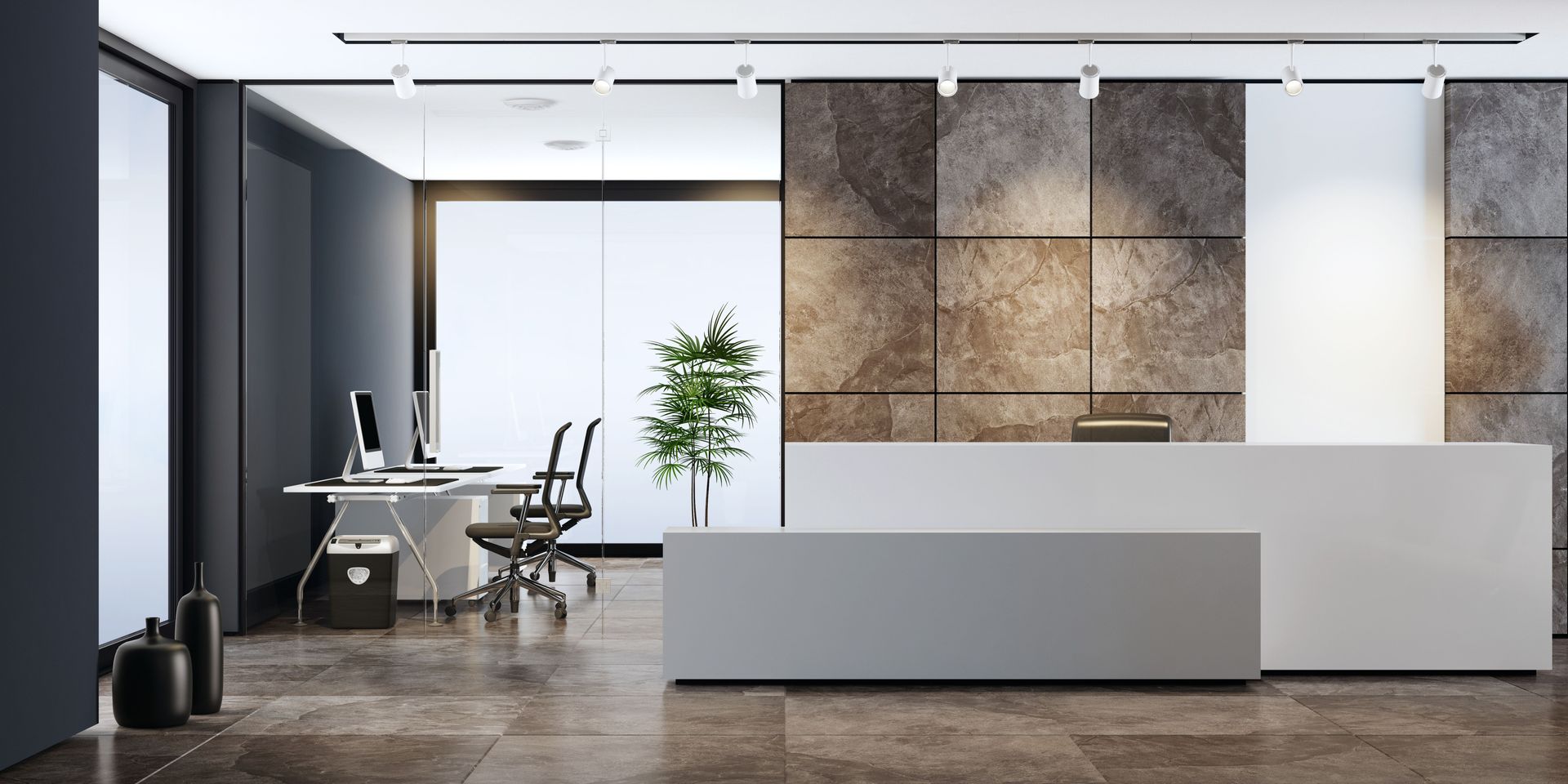 Modern office reception area with a gray and white reception desk, stone wall backdrop, and a glimpse of an office workspace through glass walls.