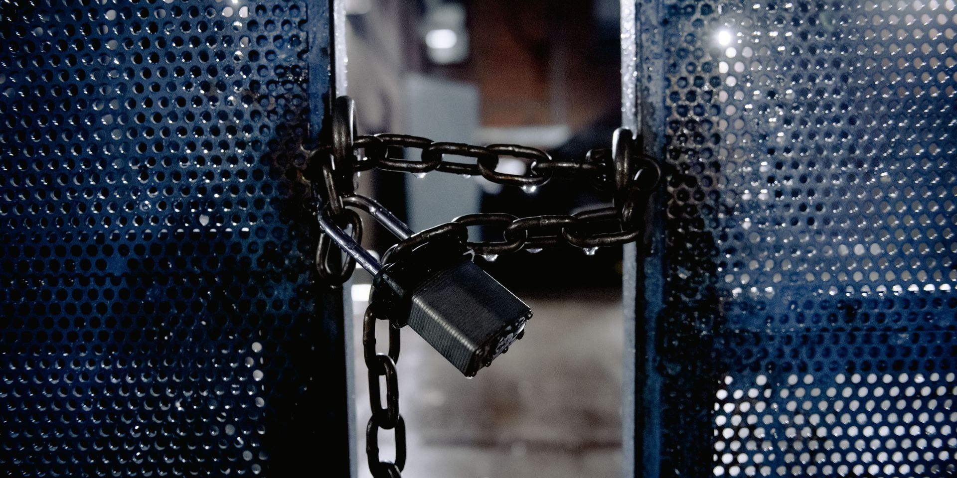 A wet, metal gate secured with a chain and padlock; droplets visible. Dark, industrial setting.